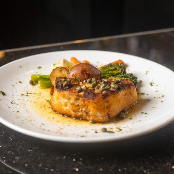 Seared fish with capers, roasted potatoes, and vegetables on a white plate at Sapore Italian Cafe.