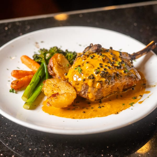 A Sapore Italian Cafe dish with a bone-in meat cut, yellow sauce, black peppercorns, and roasted vegetables.