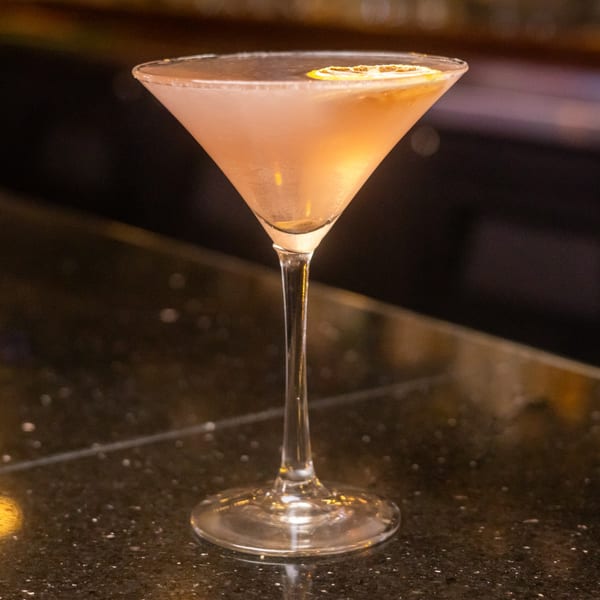 A martini glass filled with a light pink cocktail, garnished with a dried orange slice.