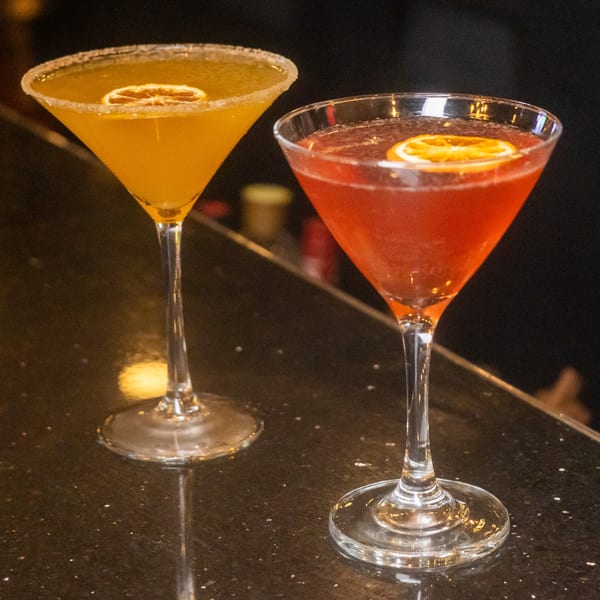 Two martini glasses, one with a yellow drink and sugared rim, the other with a red drink, on a bar.