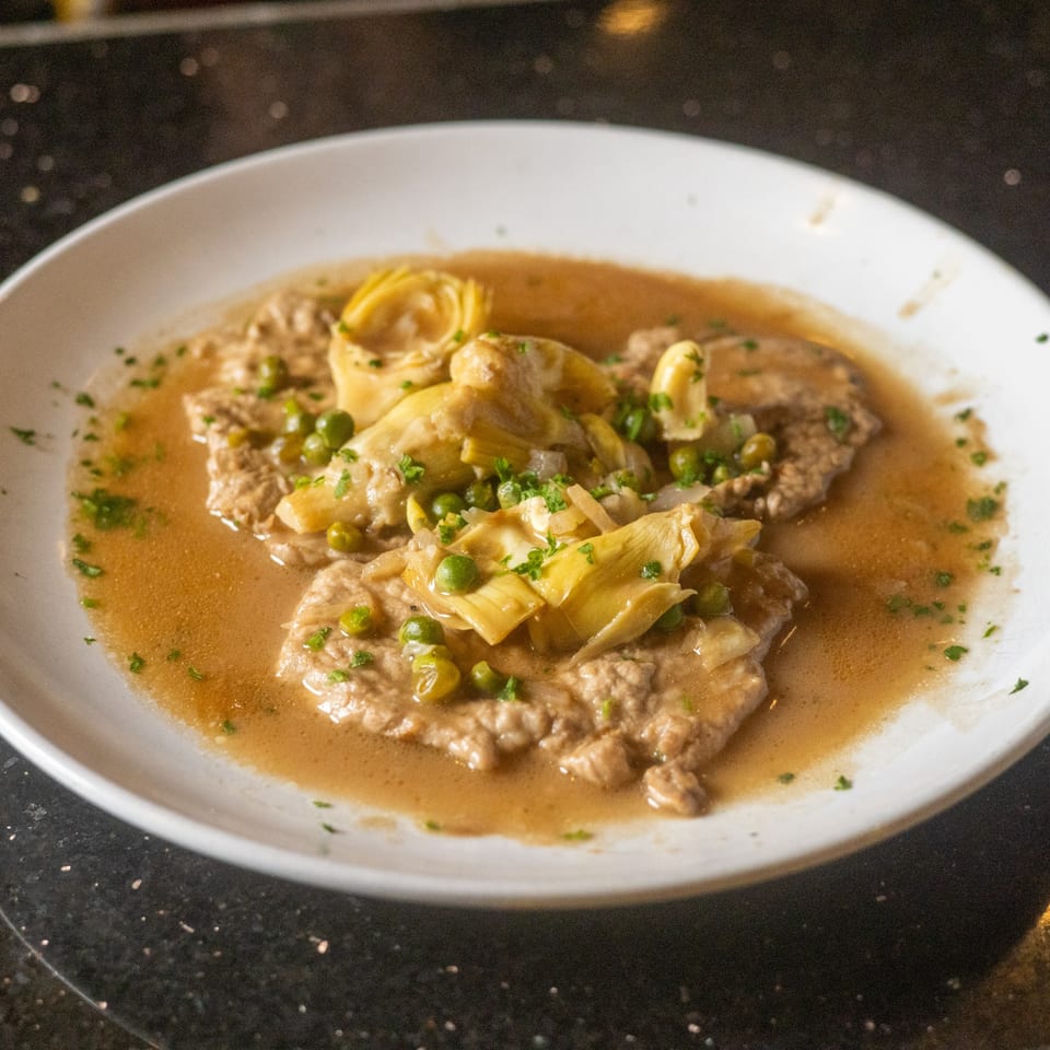 Veal scallopini with artichokes, peas, and parsley in a light brown sauce on a white plate.