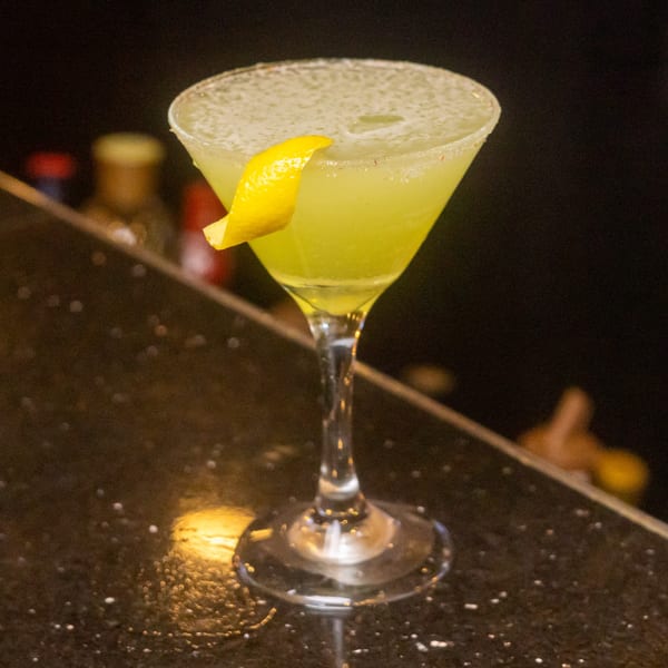 A vibrant green cocktail in a martini glass, garnished with a lemon twist, on a dark bar.
